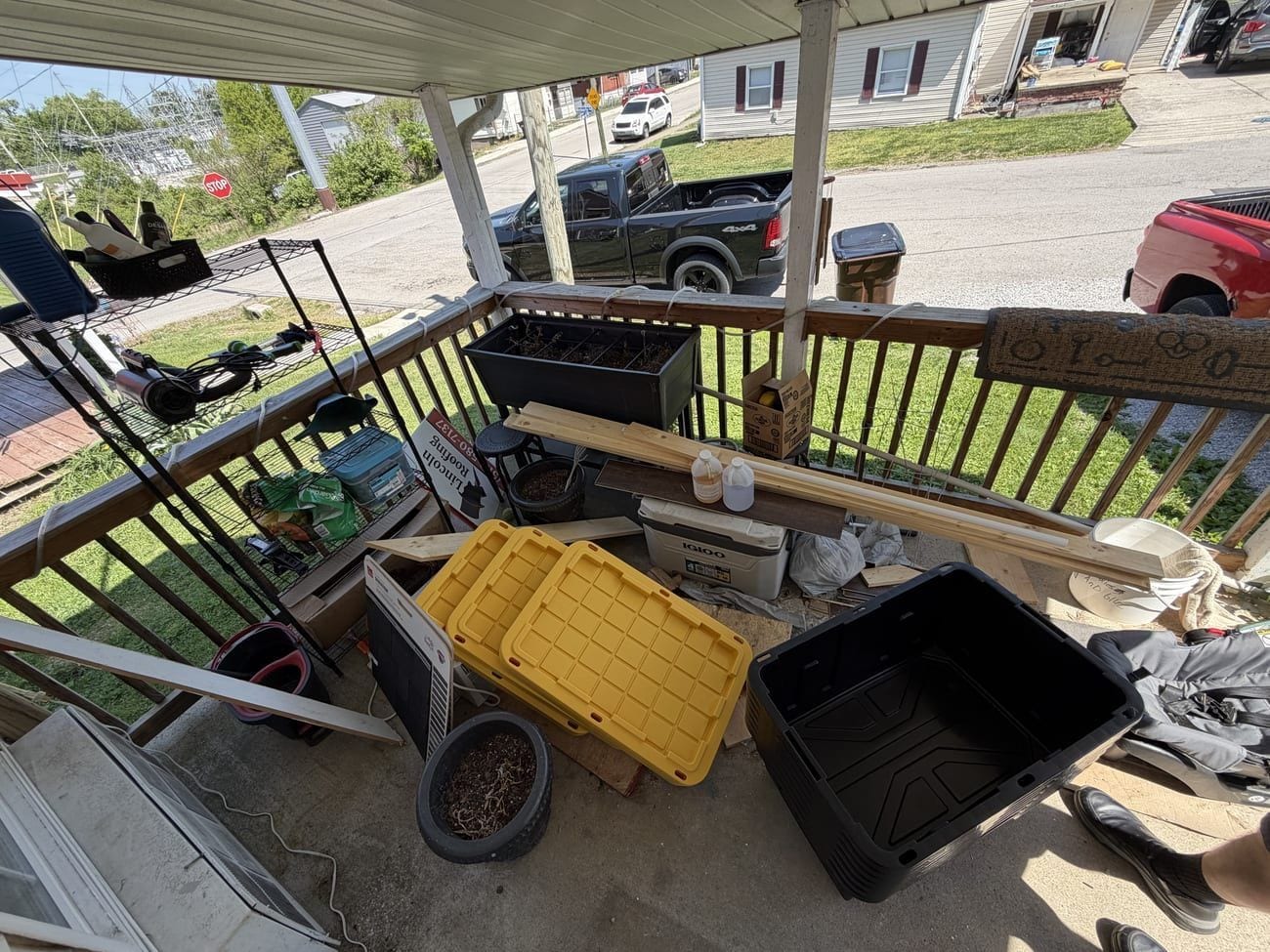 A covered front porch crowded with storage totes, planters, an appliance, and stacked lumber before a cleanout.