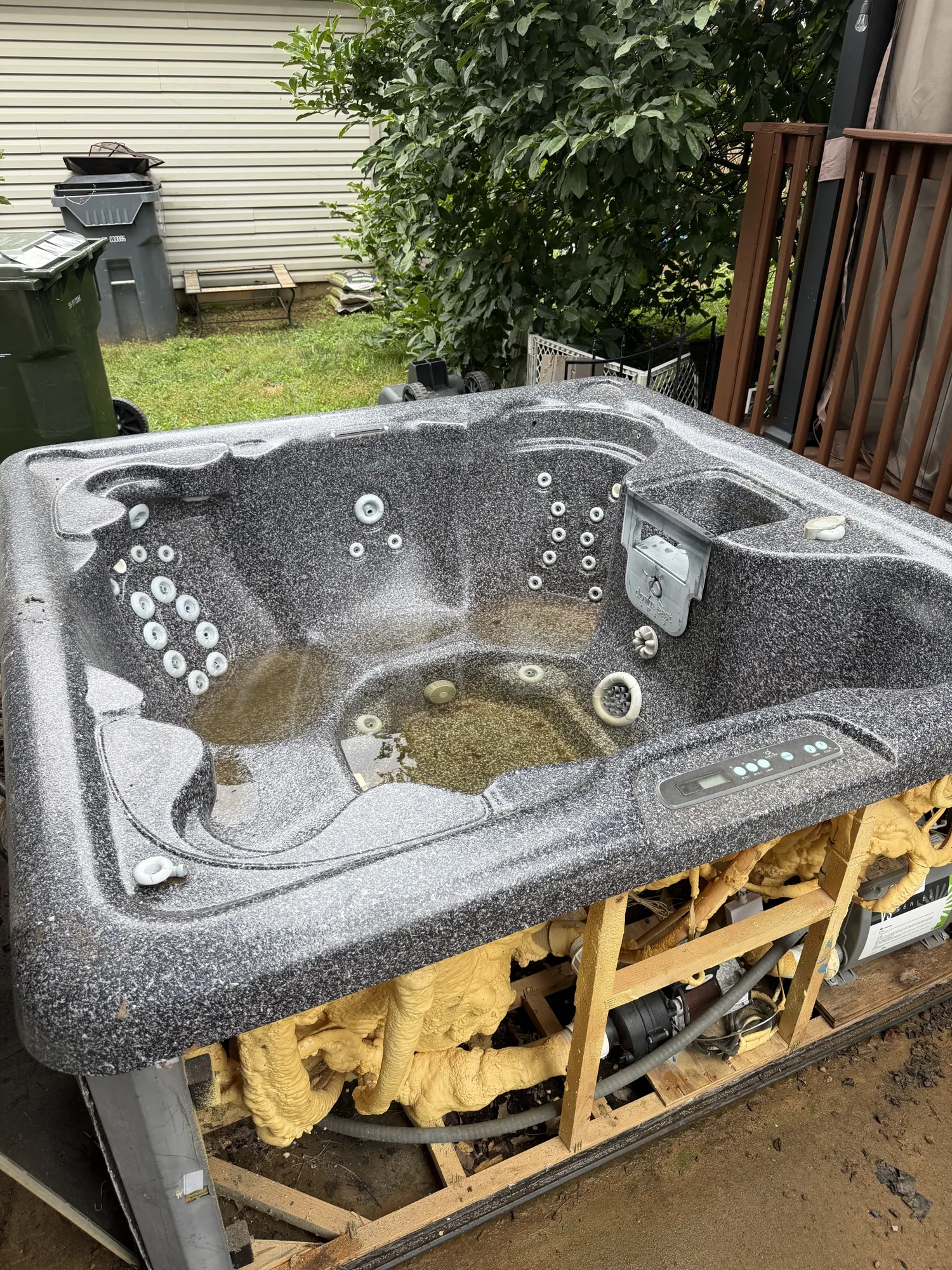 Grey speckled hot tub still sitting on the concrete pad beside the house, wheelie bins to the left and deck railing to the right.